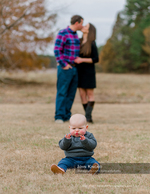 baby seated in a field with parents kissing in the background family photography